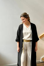 Lataa kuva Galleria-katseluun, Kuvassa seisoo alaspäin katsova nainen, jolla on säteilevä Säihke toppi päällä. Toppi on sidottu vyöllä vyötäröstä. Naisen päällä on myös vaaleat satiinihousut ja musta kangastakki.