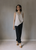 Lataa kuva Galleria-katseluun, Kuvassa seisoo nainen, jolla on päällä vaalea Säihke toppi. Toppi laskeutuu kauniisti lantioon saakka. Tämän lisäksi naisella on päällä mustat satiinipintaiset housut ja kullanhohtoiset korot. Toppi on puettu rennosti toispuoleisesti housun sisään.