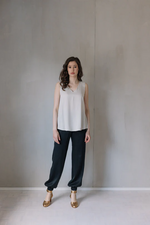 Lataa kuva Galleria-katseluun, Kuvassa seisoo nainen, jolla on päällä vaalea Säihke toppi. Toppi laskeutuu kauniisti lantioon saakka. Tämän lisäksi naisella on päällä mustat satiinipintaiset housut ja kullanhohtoiset korot.